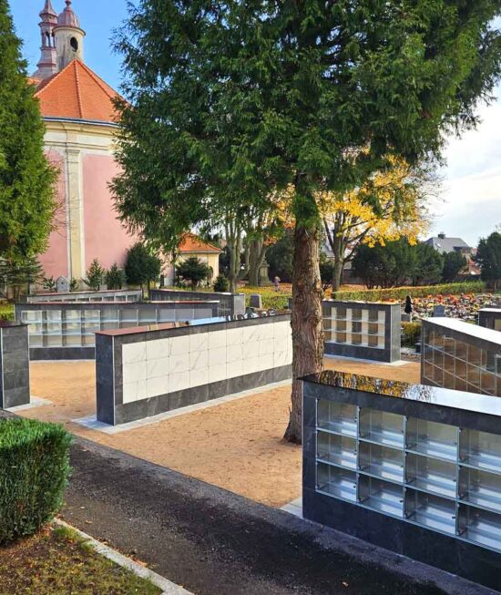 Kolumbárium na hřbitově – moderní a důstojné řešení pro uložení uren s popelem zesnulých. Tento elegantní a prostorově úsporný prvek poskytuje klidné místo pro vzpomínku a uctění památky blízkých. Umístění v upraveném hřbitovním prostředí nabízí kombinaci pietní atmosféry a praktického využití místa. Kolumbária se stávají stále častější součástí městských i venkovských hřbitovů díky své ekologičnosti, dostupnosti a estetické hodnotě. Fotografie zachycuje typickou urnovou stěnu v harmonickém souladu s okolní krajinou, ideální pro prezentaci na webu pohřební služby, kamenictví nebo městské správy.
