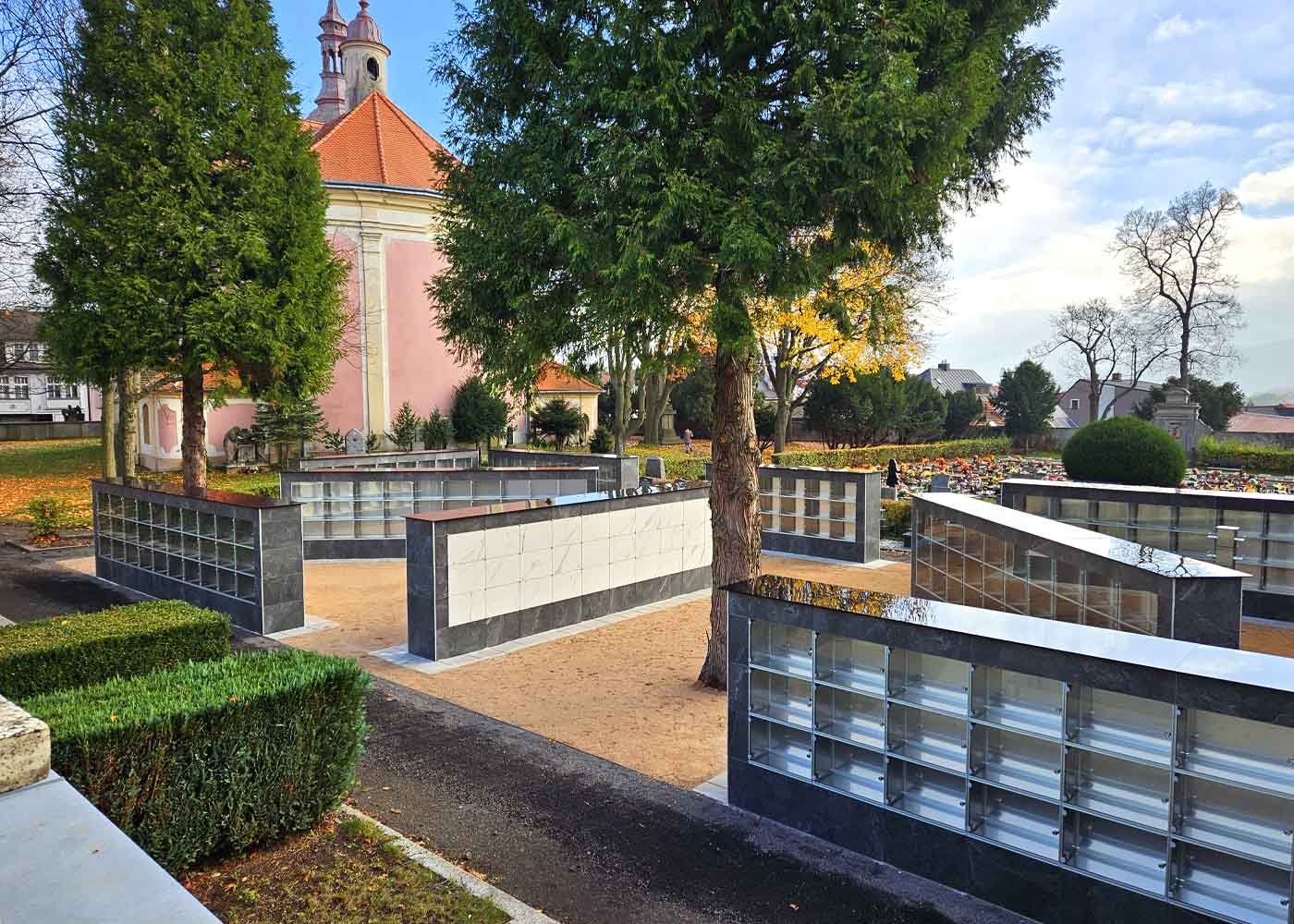 Kolumbárium na hřbitově – moderní a důstojné řešení pro uložení uren s popelem zesnulých. Tento elegantní a prostorově úsporný prvek poskytuje klidné místo pro vzpomínku a uctění památky blízkých. Umístění v upraveném hřbitovním prostředí nabízí kombinaci pietní atmosféry a praktického využití místa. Kolumbária se stávají stále častější součástí městských i venkovských hřbitovů díky své ekologičnosti, dostupnosti a estetické hodnotě. Fotografie zachycuje typickou urnovou stěnu v harmonickém souladu s okolní krajinou, ideální pro prezentaci na webu pohřební služby, kamenictví nebo městské správy.